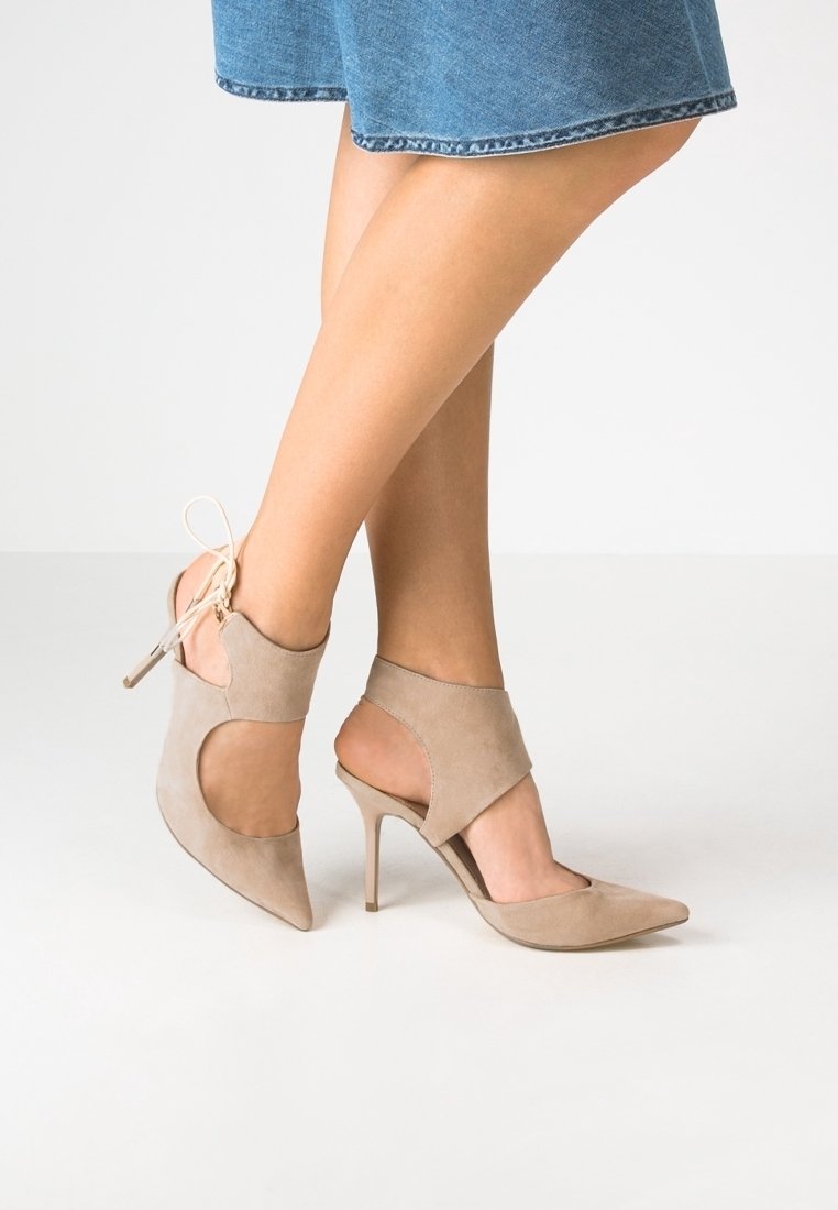 Jambes portant des chaussures à talons hauts en suède beige avec des découpes et des liens à la cheville, associées à une jupe en jean longueur genou sur un sol blanc.
