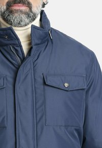 Chaqueta aislante de color azul marino con un alto cuello, cierre de cremallera y dos bolsillos frontales con botón a presión. Tejido suave con un acabado mate.