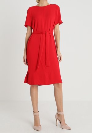 Vrouw die een rode jurk tot op de knie draagt met korte mouwen en een tailleband, gecombineerd met beige enkellaarzen met hoge hakken en een enkelriem.