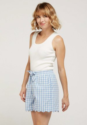 Femme blonde aux cheveux ondulés portant un haut blanc sans manches et un short vichy bleu à ourlet festonné et taille nouée, debout devant un fond neutre.