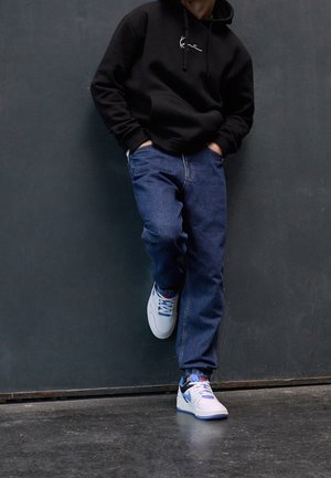 Person lehnt an einer dunklen Wand, trägt einen schwarzen Hoodie, blaue Jeans und weiße Nike-Sneaker mit schwarzem Swoosh und blauen Akzenten.