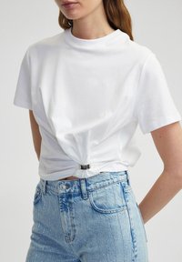 Witte katoenen t-shirt met een ronde hals en korte mouwen, voorzien van een geknoopt detail bij de taille met een zilveren hardware accent, gecombineerd met lichtblauwe spijkerbroeken.