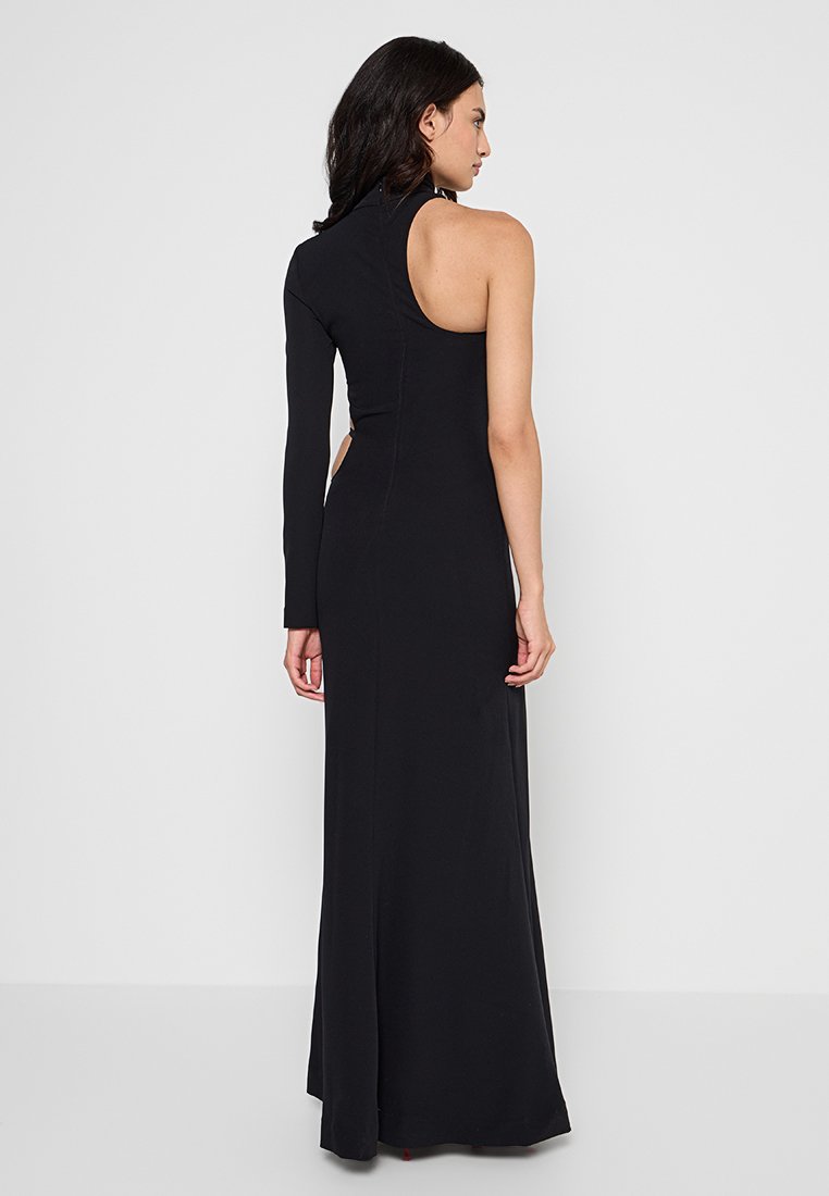 Femme portant une robe noire longue avec une manche longue sur le bras gauche et l'épaule droite découverte, se tenant devant un fond uni.