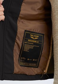 Black softshell jacket with a brown interior. The label lists features and specifications in yellow text, including fabric details and design elements.