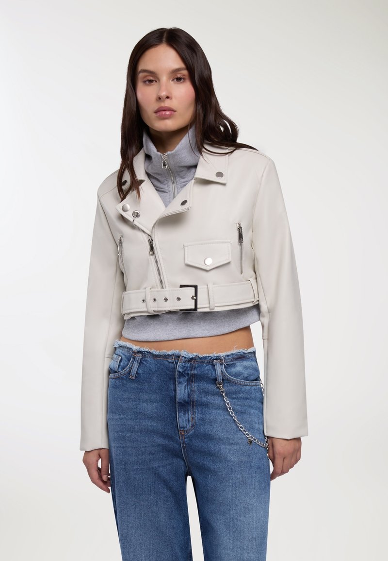 Giacca corta in pelle bianca con accessori in argento, tasche con cerniera e cintura. Indossata sopra una maglia grigia a collo alto e abbinata a jeans di denim blu.
