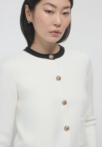 Weißer Cardigan mit schwarzem Kragen, ausgestattet mit fünf goldfarbenen Knöpfen und zwei Fronttaschen, aus weichem Strickmaterial mit strukturierter Oberfläche.