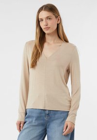 Beige Langarmshirt mit V-Ausschnitt, aus weichem Stoff gefertigt. Es hat ein tailliertes Design und eine dezente Struktur, kombiniert mit blauen Jeans.
