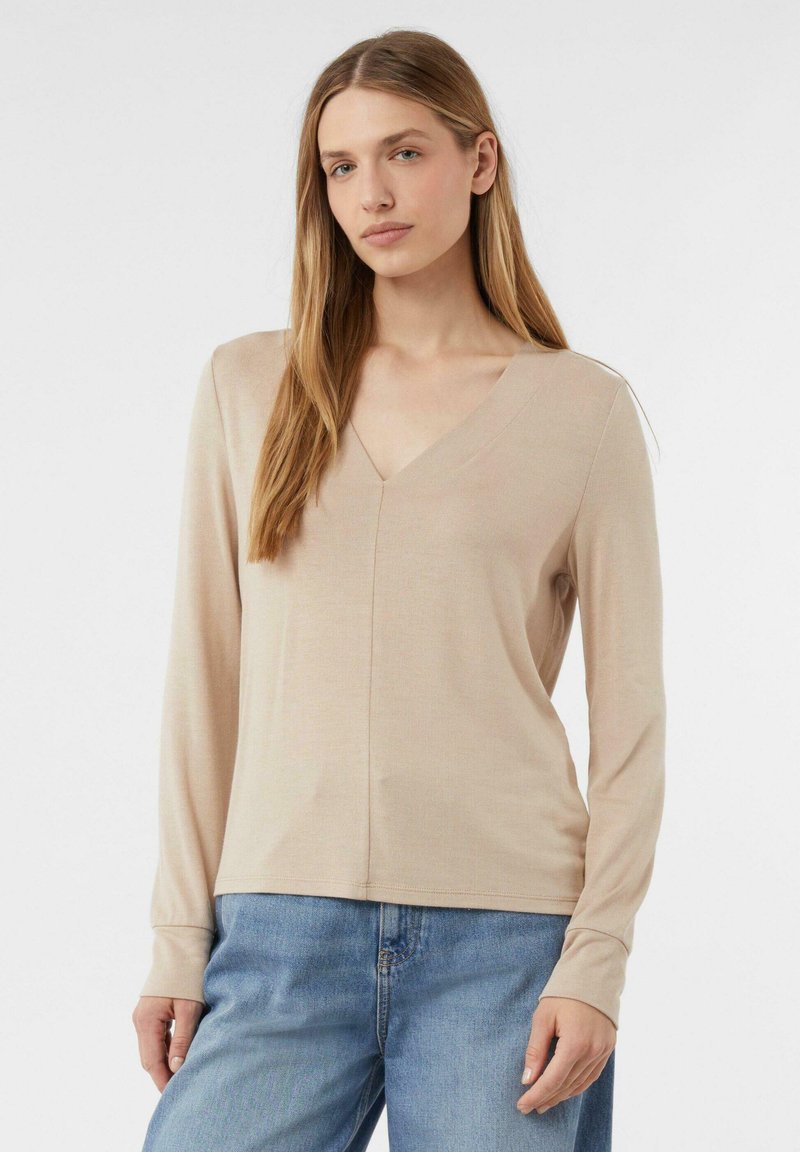 Beige Langarmshirt mit V-Ausschnitt, aus weichem Stoff gefertigt. Es hat ein tailliertes Design und eine dezente Struktur, kombiniert mit blauen Jeans.