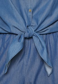 Le tissu en denim bleu présente un détail de taille nouée avec une texture lisse. Des boutons à finition dorée sont présents sur le devant.