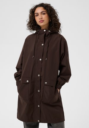 Young woman with curly hair wearing a dark brown hooded raincoat with snap buttons and large front pockets, hands in pockets, neutral background.