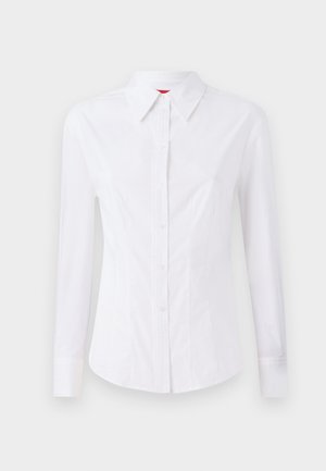 Hvit langermet skjorte med spiss krage, frontlukking med knapper og figurnær design. Glatt bomullsstoff med subtile søm detaljer.