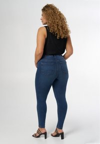 Jean skinny en denim bleu taille haute avec une texture lisse et deux poches arrière, porté avec un top sans manches noir et des sandales à talons noires.