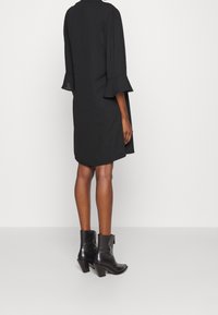Robe noire à manches courtes avec des poignets volantés, présentant un design fluide. Portée avec des bottines en cuir noir et une silhouette texturée et structurée.