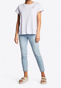 T-shirt bianco in cotone con maniche corte arrotolate e un piccolo design di fenicottero viola. Abbinato a jeans attillati azzurri e sandali bianchi.
