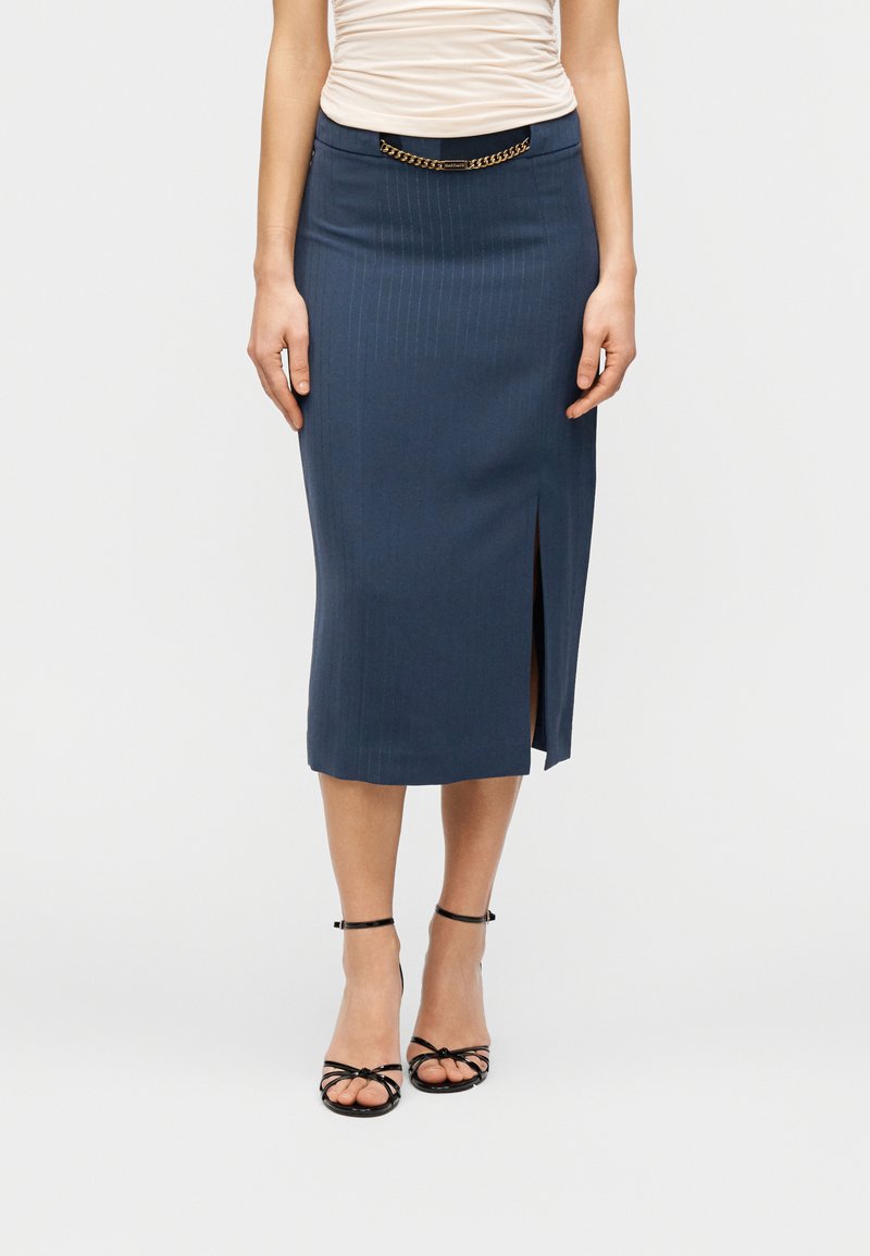 Εξατομικευμένη μολυβοειδής φούστα σε ναυτικό μπλε με κάθετες λεπτές ρίγες, πλάγια σκίσιμο και μαύρη ζώνη μέσης με χρυσή λεπτομέρεια αλυσίδας. Συνδυασμένη με μαύρα πέδιλα με λεπτά λουράκια.