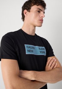 Juodas marškinėliai su mėlynu stačiakampiu grafiku, užrašu "CALVIN NEW KLEIN YORK". Trumpomis rankovėmis ir apvaliu kaklu. Lygus audinys.