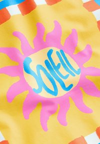 Textil s žlutým pozadím a velkým růžovým slunečním vzorem, modrým textem "SOLEIL" uprostřed a barevnými kostkovanými doplňky.