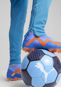 Zili sporta bikses ar rāvējslēdzēja piedurknēm, kas saskaņotas ar ziliem un oranžiem futbolbotiem uz zila un melna rakstaina futbolbala.