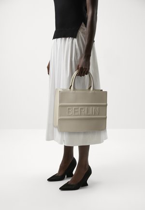 Personne en haut noir et jupe blanche tenant un sac beige avec l'inscription "BERLIN" embossée, portant des talons noirs à bouts pointus, debout sur un sol blanc.