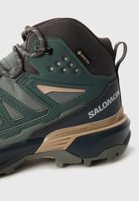 Žalios ir pilkos žygio bateliai su tinklo ir sintetinės medžiagos viršutine dalimi, Gore-Tex etiketė, guminis padas ir detali apdaila šonuose bei kulne.