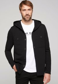 Giacca nera con cappuccio, bottoni, due tasche sul petto e un tessuto texturizzato, indossata sopra una T-shirt bianca con testo stampato.