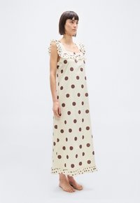 Γυναίκα που στέκεται ξυπόλητη σε μάξι φόρεμα κρεμ χωρίς μανίκια, με μεγάλα καφέ πουά και βολάν στους ώμους και στο τελείωμα.