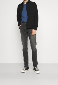 Chaqueta negra sobre un polo azul, combinada con jeans grises y zapatillas negras. Texturas suaves, corte ajustado y detalles sutiles en la chaqueta.