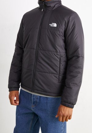 Homme portant une veste matelassée noire North Face avec une fermeture éclair à l'avant et un jean bleu sur un fond uni.