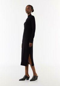 Schwarzes Langarmkleid mit hohem Kragen, seitlichen Schlitzen und taillierter Silhouette. Kombiniert mit schwarzen hohen Mules. Glatte Textur, minimalistisches Design.