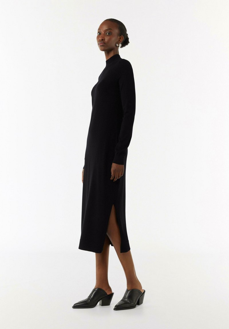 Schwarzes Langarmkleid mit hohem Kragen, seitlichen Schlitzen und taillierter Silhouette. Kombiniert mit schwarzen hohen Mules. Glatte Textur, minimalistisches Design.