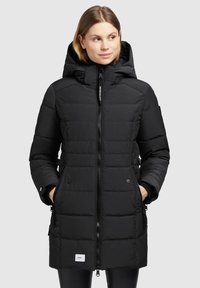 Kvindelig iført en lang sort quilted vinterfrakke med hætte, frontlynlås, sidelommer, og hænderne i lommerne, vendende let mod højre.