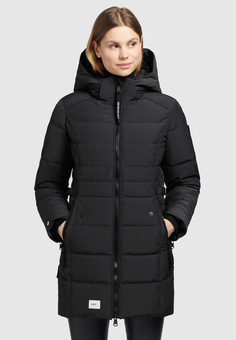 Kvindelig iført en lang sort quilted vinterfrakke med hætte, frontlynlås, sidelommer, og hænderne i lommerne, vendende let mod højre.