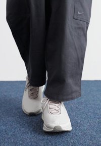 Sneakers Nike di colore grigio chiaro con un motivo testurizzato, abbinati a pantaloni neri larghi con tasca laterale e un sottile dettaglio del logo.