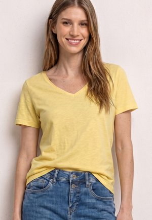 Jeune femme souriante, portant un T-shirt jaune à col en V et un jean bleu, debout devant un mur clair.