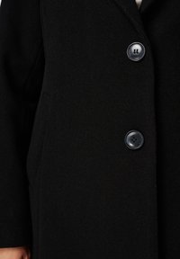 Manteau en laine noire avec deux grands boutons foncés fixés à l'avant, montrant une partie d'une poche et une main sur le côté.