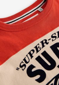 Červená a krémová mikina s okrúhlym výstrihom. Obsahuje veľký námornícky text s nápisom "SUPER" a detaily v textúrovanej tkanine. Tag je viditeľný vo vnútri výstrihu.