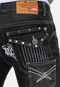 Black denim jeans close-up showing back pocket with white stitching, metal studs, button, and a tan leather brand label reading Kosmo Lupo 1996.
