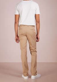 Béžové chinos kalhoty s úzkým střihem, z bavlněného materiálu, se dvěma zadními kapsami, nošené s bílým tričkem a bílými teniskami, zobrazené zezadu.