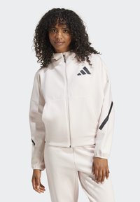Lichtroze zip-up jack met een hoge kraag, grote voorzakken en zwarte logo-accenten op de borst en mouwen. Gladde stoftextuur.