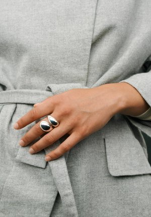 Hand ruht auf einem hellgrauen Mantelgürtel und trägt einen modernen silbernen Doppelbandring am Mittelfinger.