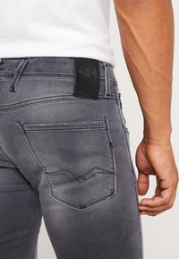 Jeans de mezclilla gris con un corte ajustado, que cuentan con un parche de cuero negro en la cintura, bolsillos traseros con costuras curvas y una textura suave.