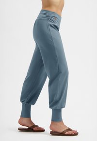 Frau in blauen, elastischen Jogginghosen mit Bündchen an den Knöcheln und braunen Flip-Flops, die vor einem schlichten weißen Hintergrund steht.