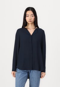 Marineblå langermet bluse med V-hals og løs passform, laget av glatt stoff. Kombinert med lyseblå jeans med sideslisser.