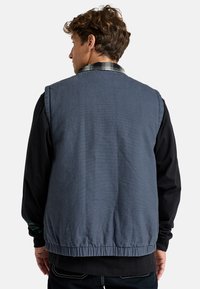 Sleeveless blue vest made of textured fabric with elastic hem, worn over a black long-sleeve shirt. Features a plaid collar detail.
