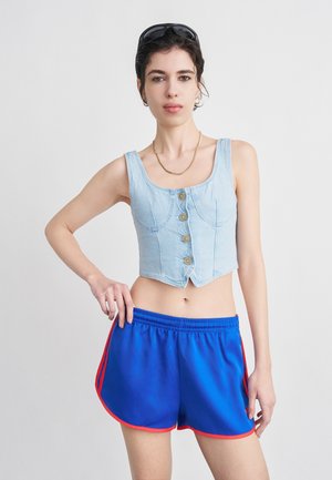 Kobieta ubrana w jasnoniebieską, rozpinaną jeansową bluzkę typu crop top i niebieskie sportowe szorty z czerwonym wykończeniem, stojąca na tle jednolitego białego tła.