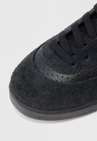 Zapatilla de ante negro con acentos de cuero, superficie texturada, cordones negros y detalles perforados en la zona del dedo del pie, con una suela de goma.