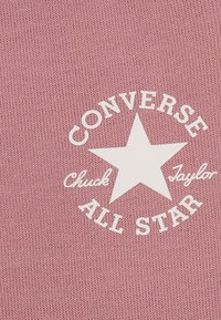Tissu rose avec une impression blanche en caoutchouc représentant une étoile et le texte "Converse Chuck Taylor All Star" en disposition courbée.