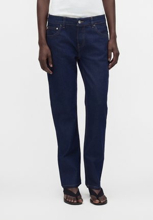 Persona che indossa jeans blu scuro a gamba dritta, top bianco e sandali neri con cinturini incrociati, in piedi su sfondo bianco.