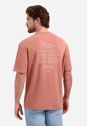 Man met bruin haar die een zalmroze t-shirt draagt met witte tekst op de rug en lichtblauwe spijkerbroek, staand met de rug naar ons toe.