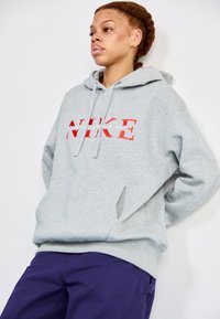 Pilkas pūkinis džemperis su kengūros kišene, išsiskiriantis ryškiu raudonu "NIKE" logotipu. Minkštas paviršius ir laisvas siluetas.